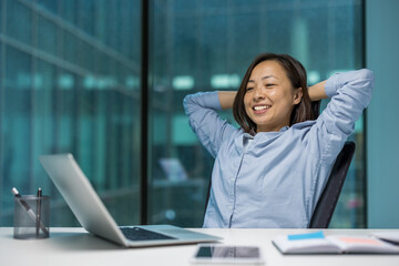 Asian businesswoman smiling, relaxing in modern office with hands behind head, laptop on desk....