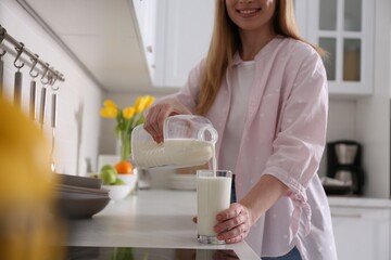 Young woman pouring milk from gallon bottle into glass at light countertop in kitchen, closeup