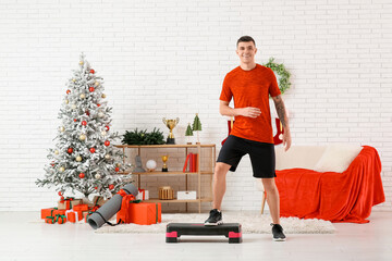 Young happy man training with aerobic stepper in living room decorated for Christmas