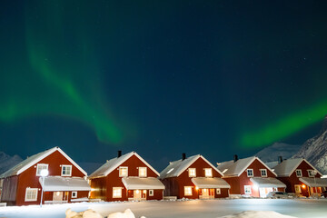 Northern lights over rorbu cabins in Svolvaer on Lofoten islands, Norway. Winter night with Aurora borealis, traditional red wooden houses, snowy mountains and starry sky. Landscape with polar lights