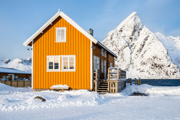 Traditional yellow rorbu house and Olstind mountain peak in Sakrisoy fishing village on Lofoten islands, Norway. Winter landscape with snowy mountains and Scandinavian cottage