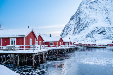 Reine village with traditional rorbu houses on Lofoten islands in winter. Scandinavian landscape with red wooden fishermen cabins on frozen sea water and snowy Reinebringen mountain in northern Norway