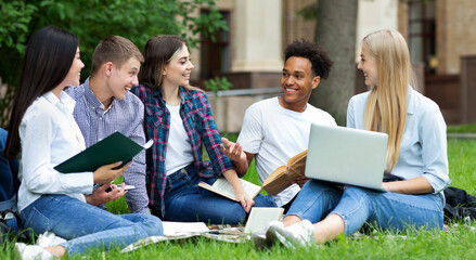 Excited students resting in campus outdoors, sitting on grass, chatting and using laptop
