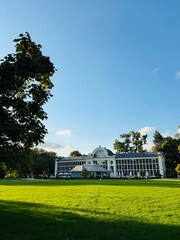 Obraz premium A bright, sunny day in a large park with a historic white building in the background. The lush green lawn stretches toward the structure, while trees frame the scene under a clear blue sky.