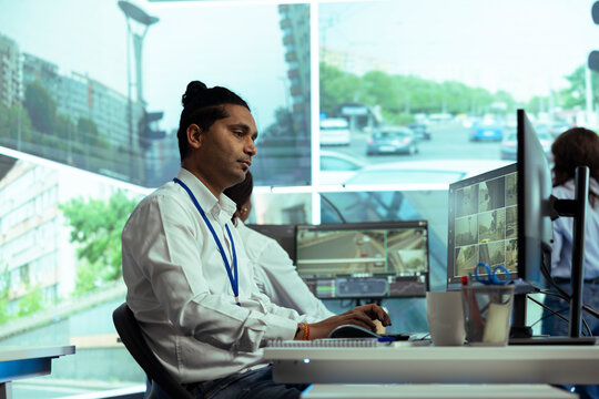 Indian man works on monitoring real time surveillance footage from traffic on CCTV cameras, reporting any illegal activity. Young employee in security control center, public safety.