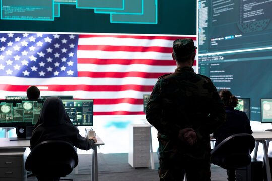 US army official working on ensuring servers protection and state secrets, protecting important hybrid warfare data in high tech agency office. Military soldier supervises hacking activity.