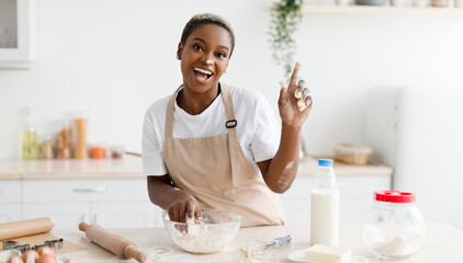 Laughing millennial african american woman in apron prepares dough for baking and raises finger up in light scandinavian kitchen interior. Got idea, cooking pizza, pie at home and facial expression