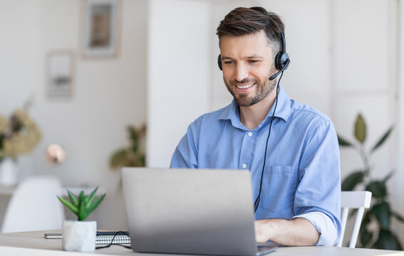 Call center operator at work. Positive male manager wearing headset and typing on laptop at workplace in modern office, consulting clients