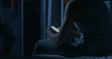 A man uses a mobile phone while sitting on the bed at night. Close-up of his hands, an unrecognizable person. Dark room, home interior