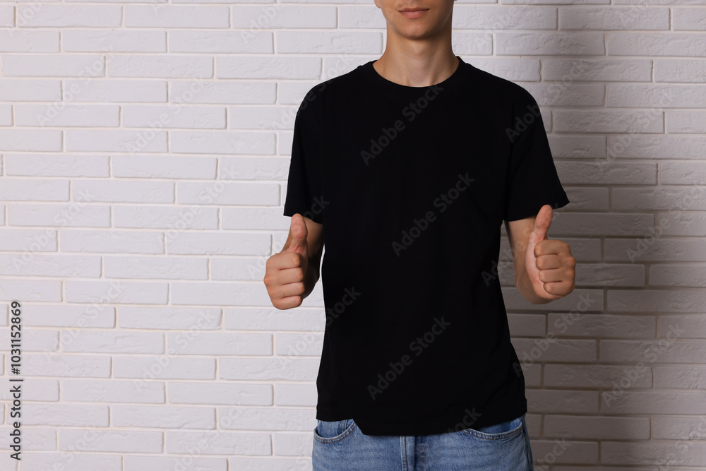 Poster Teenage boy wearing black t-shirt and showing thumbs up near white brick wall, closeup. Space for text