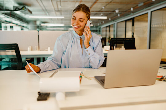 A young businesswoman on the phone at her desk, analyzing documents and using her laptop effectively