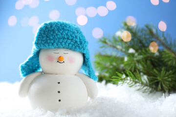 Cute decorative snowman and fir branches on artificial snow against blurred lights, closeup