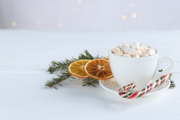 Yummy cocoa with marshmallows in cup, candy cane, dried orange slices and fir tree branches on white wooden table. Space for text