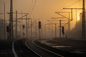 The train platform glows softly in the morning light, with dawn's golden hue wrapped in mist. A red train slowly arrives, ready for the early crowd.