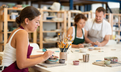 During workshop at master class, female employee paints clay vessel, applying pigment to plate with...