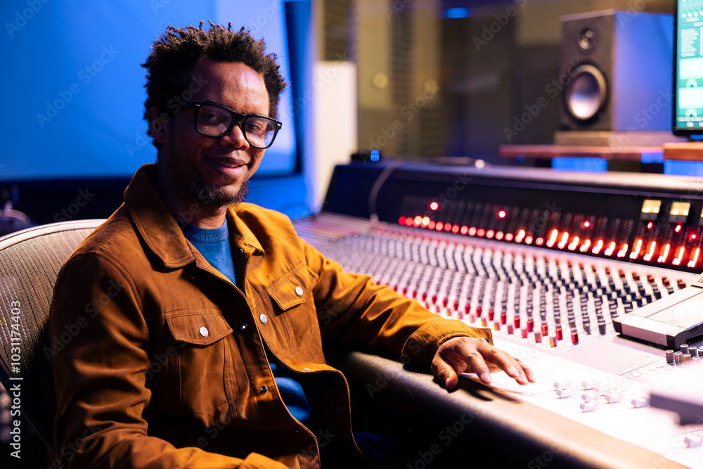 Poster Portrait of african american sound engineer producing records in studio, working with digital audio software and professional mixing console in control room. Expert adjusting volume levels on songs.