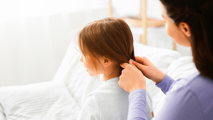 Fototapeta premium Mother care concept. Close up of woman braiding her little daughter hair, home interior