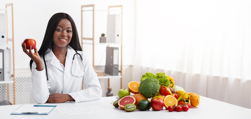 African american smiling female doctor dietologist sitting at workdesk, holding apple in her hand, empty space