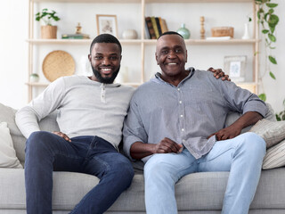 Portrait of adult black son and his senior father sitting on couch at home, posing to camera, free...