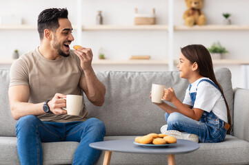 Snack Time. Portrait of cheerful little girl and man holding cups, drinking coffee and tea, eating cookies and looking at each other, dad and daughter talking sitting on the couch in living room