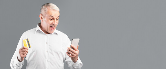 Unbelievable. Excited surprised senior man holding credit card and smartphone, gray studio wall, free space