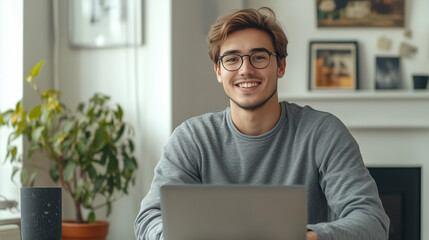 A young man sitting at the table with his laptop, smiling and looking into camera against white wall.