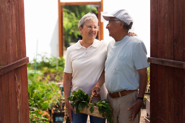 A couple is standing in front of a greenhouse, holding a basket of vegetables. The man is wearing a gray cap and the woman a white shirt.