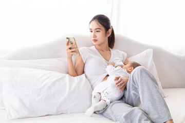 Asian Young mother holding her newborn child and breastfeeds baby while her talking on the phone, busy with call on bed. Adorable infant sucking a woman breast after wake up in morning.