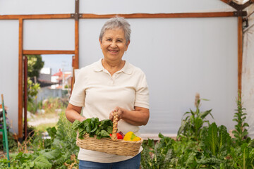 A woman is holding a basket of vegetables and smiling. She is in a greenhouse, surrounded by plants