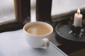 A warm cup of coffee rests beside an open book on a windowsill, while a lit candle flickers softly, creating a serene atmosphere against the snowy backdrop.