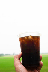 Cropped hand holding plastic cup of iced Americano coffee against green rice fields background at countryside of Thailand