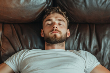 Fototapeta premium A bearded man in a white t-shirt rests peacefully with his eyes closed, lying on a brown leather couch