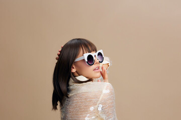 Beautiful young woman wearing sunglasses holds a seashell against a beige background, exuding a relaxed summer vibe and beach aesthetics in a minimalist, stylish setting