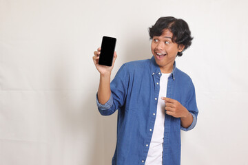 Portrait of excited Asian man in blue shirt showing and presenting blank screen mobile phone for...