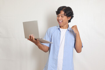 Portrait of excited Asian man in blue shirt holding laptop and showing happy face expression. Isolated image on white background