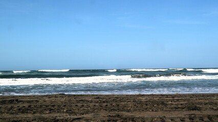 Waves crashing against the shore with a clear blue sky in the background at Parangtritis Beach, Yogyakarta.