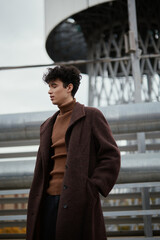Fashionable man in brown coat and turtleneck sweater standing in front of tall building with water...