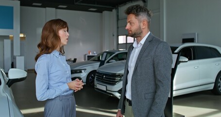 Fototapeta premium A car dealership manager in formal clothes smiles and talks to a customer standing near cars in a car dealership