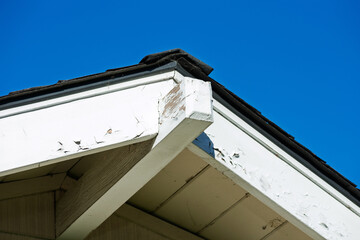 Roof eaves with chipped and peeling white paint, revealing wear and tear against a clear blue sky