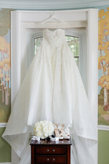 Bridal dress and wedding accessories in the form of shoes, rings and a bouquet. White wedding dress is hung near the window. Beautiful bridal bouquet of white roses. High-heeled shoes and rings