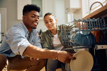 Happy multiracial couple using dishwasher in  kitchen.