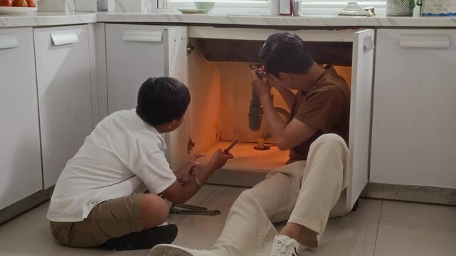 Long shot of teenage Asian son and his father stopping leak in pipe under sink at home kitchen