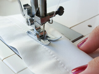 A woman sews on a sewing machine. Hands sew white fabric on a sewing machine. White line sewing stitch on white fabric. Sewing production, tailoring, atelier.