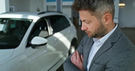 A man inspects a car standing in a car dealership with his arms folded on his chest with a serious look