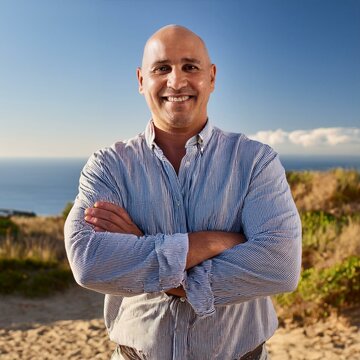 Un hombre calvo, de aproximadamente 40 a&ntilde;os, con una sonrisa amplia y una expresi&oacute;n de confianza. Est&aacute; al aire libre, en un parque verde y soleado, con el cielo azul de fondo.