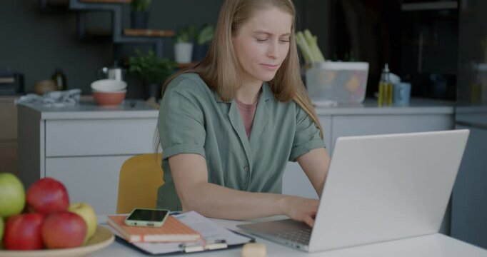 Young lady freelance worker using laptop typing sitting in kitchen at table concentrated on online activity. Freelance job and domestic workplace concept.