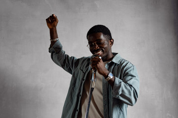 Handsome young African American singer standing with retro microphone singing song on grey...