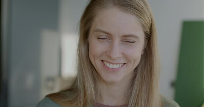 Close-up slow motion portrait of cheerful young woman blonde standing indoors in sunlit home smiling looking at camera. People and good mood concept.