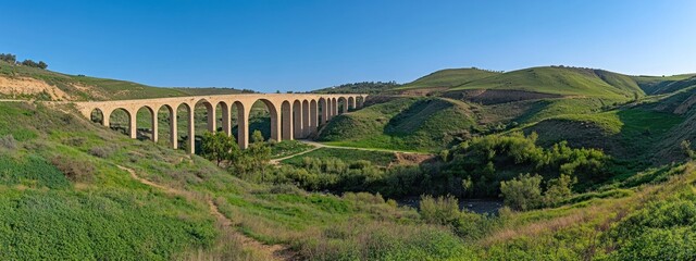 A stone arched bridge stretches across a valley with green hills and a clear blue sky.