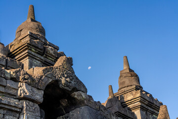 Sunset light on ancient reliefs, stupa and Buddha statues in niche with moon in background between stupas, at the ancient historical Buddhist UNESCO site of Borobudur (Barabudur), Magelang, Indonesia.
